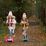 Two girls wearing beanies and winter jackets ride scooters on a leaf-strewn path in an autumn park.