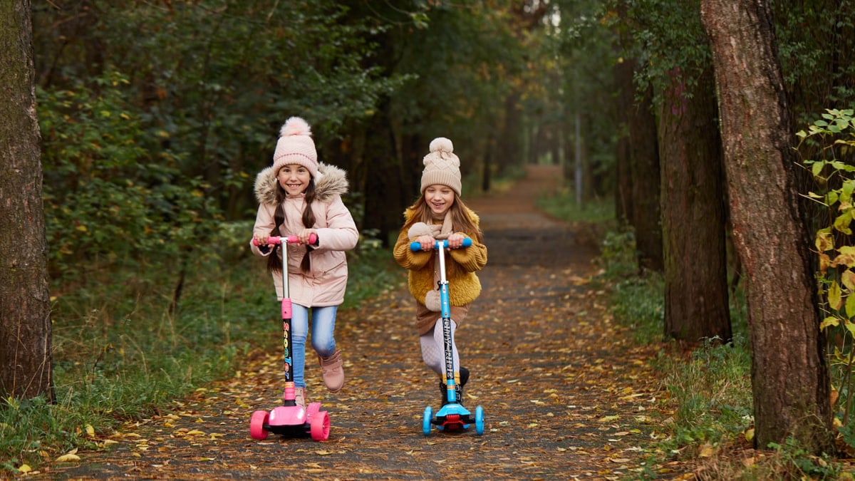 Two girls wearing beanies and winter jackets ride scooters on a leaf-strewn path in an autumn park.